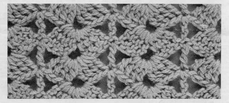 May include: Close-up of a crocheted fabric with a repeating pattern of small flowers. The fabric is a light gray color and has a textured surface.