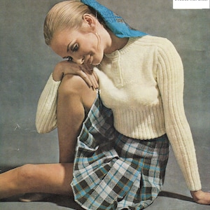 May include: A woman wearing a white sweater and a plaid skirt. She is sitting with her head down and her hands on her knees. The sweater is ribbed and the skirt is a blue, black, and white plaid. The text "DK yarn 34/36/38 inch chest" is in the top right corner of the image.