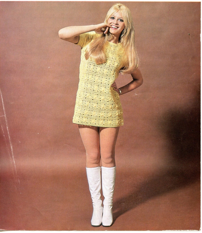 May include: A woman wearing a yellow crochet mini dress and white knee-high boots. She is standing in front of a brown background.
