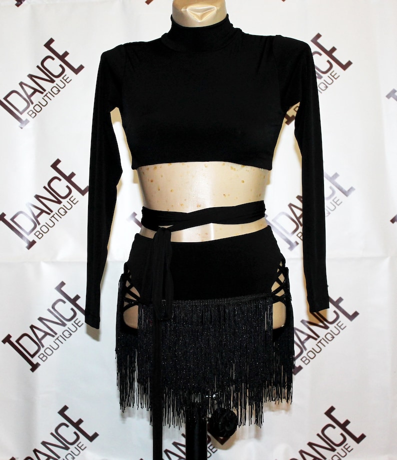 May include: Black dance costume comprising a long-sleeved crop top and a skirt with fringe and cut-out details. The skirt has a wrap-around tie. The costume is displayed on a mannequin.