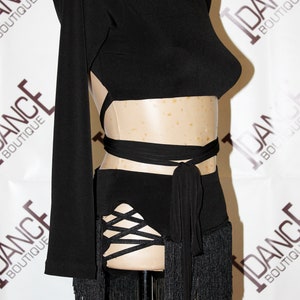 May include: Black dance costume with a high neck, long sleeves, and open back design. The costume features a wrap-around waist tie and a skirt with fringe and crisscross detailing. The words "DANCE BOUTIQUE" are visible in the background.