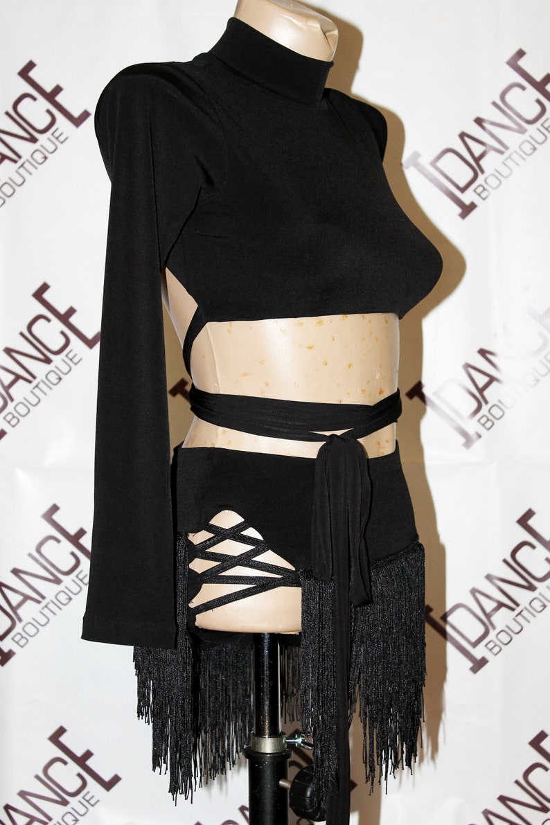 May include: Black dance costume with a high neck, long sleeves, and open back design. The costume features a wrap-around waist tie and a skirt with fringe and crisscross detailing. The words "DANCE BOUTIQUE" are visible in the background.