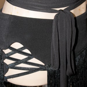 May include: Black dance belt with a wrap-around tie and fringe detailing. The belt features a crisscross design on the side and is made of a soft, flowing fabric. The fringe adds movement and flair to the garment.