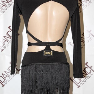 May include: Black dance costume with long sleeves and a high neckline. The back features a large cutout and crisscross straps. The skirt has a fringe detail. The IDANCE logo is visible on the back. The costume is displayed on a mannequin.