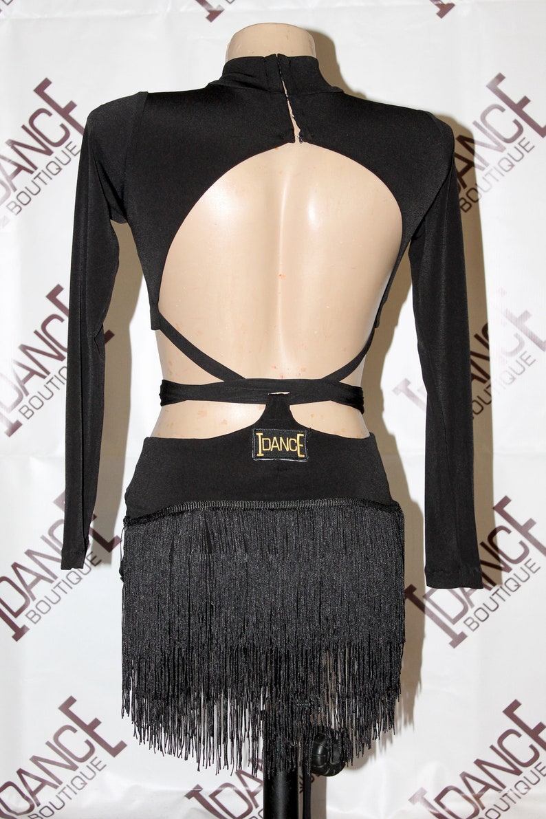 May include: Black dance costume with long sleeves and a high neckline. The back features a large cutout and crisscross straps. The skirt has a fringe detail. The IDANCE logo is visible on the back. The costume is displayed on a mannequin.