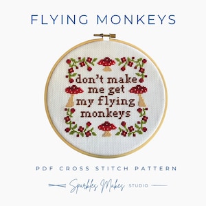 May include: A cross-stitch embroidery hoop with the text "don't make me get my flying monkeys." The design includes red mushrooms and floral accents. The words "FLYING MONKEYS" and "PDF CROSS STITCH PATTERN" are above and below the embroidery. The artist's name is "Sparkles Makes Studio."