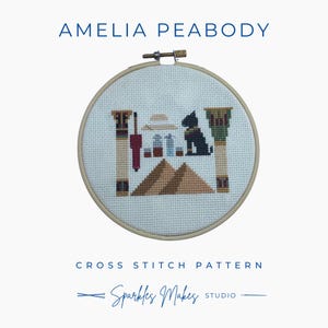 Op de afbeelding: Een kruissteekpatroon met een Egyptisch thema, met piramides, zuilen en een zwarte hond. Het ontwerp is omlijst in een houten hoepel, met de tekst "AMELIA PEABODY" en "CROSS STITCH PATTERN". De studio naam "Sparkles Makes" is ook zichtbaar.