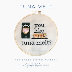 May include: A cross-stitch design in a wooden hoop, with the words "TUNA MELT" above a ginger ale can and a tuna melt sandwich. The text "you like tuna melt?" is below the sandwich. The words "PDF CROSS STITCH PATTERN" and "Sparkles Makes Studio" are also included.
