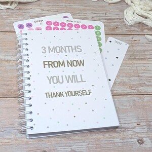 A weight loss journal with planner stickers and bookmark on a wooden desk, the journal cover states "3 months from now you will thank yourself". The stickers have activity, exercise symbols and also ticks and crosses.