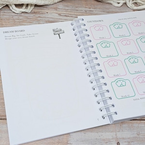 Puede incluir: Una agenda de espiral abierta en una p&aacute;gina titulada DREAM BOARD con instrucciones para establecer metas. La p&aacute;gina opuesta presenta una secci&oacute;n COUNTDOWN con una cuadr&iacute;cula de iconos de b&aacute;scula etiquetados de Week 1 a Week 10 en tinta rosa y verde.