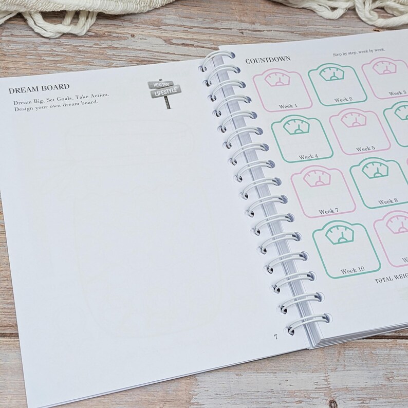 Puede incluir: Una agenda de espiral abierta en una p&aacute;gina titulada DREAM BOARD con instrucciones para establecer metas. La p&aacute;gina opuesta presenta una secci&oacute;n COUNTDOWN con una cuadr&iacute;cula de iconos de b&aacute;scula etiquetados de Week 1 a Week 10 en tinta rosa y verde.