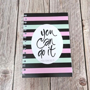 May include: A black and white striped notebook with a pink and green stripe pattern. A white circle with the text "you can do it" is on the front cover. The notebook has a spiral binding.
