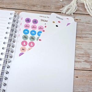 Puede incluir: Una agenda de espiral blanca con un bolsillo que contiene una hoja de pegatinas con iconos circulares de colores que representan diversas actividades f&iacute;sicas, como correr, nadar y ciclismo, junto a una pesta&ntilde;a blanca con la palabra TODAY.