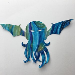 Op de afbeelding: Een blauwe en groene stoffen uitsnijding van een Cthulhu-monster met tentakels en vleugels.