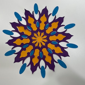 May include: A purple paper mandala with a yellow center and blue accents. The mandala is made of paper cutouts and has a symmetrical design.