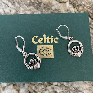 May include: Silver-toned Claddagh earrings with lever-back closures. Each earring features a heart, crown, and hands design within a circular frame. The earrings rest on a green box with the word "Celtic" in gold lettering.