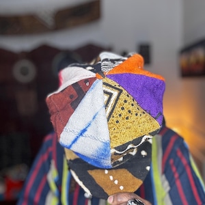 May include: A colorful patchwork hat with geometric patterns in various colors, including orange, purple, and yellow. The hat is made of different fabric pieces sewn together. A silver ring with a shell design is visible.