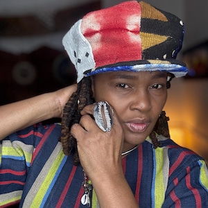 May include: A colorful patchwork hat with red, white, black, and gold sections. The hat has a flat brim and is worn with a striped shirt featuring blue, green, yellow, and red stripes. A large silver ring in the shape of a shell is also visible.