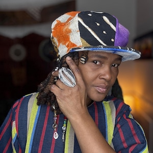 May include: A colorful patchwork baseball cap with orange, black, purple, and white patterns. The cap has a blue brim and is worn with a striped shirt in blue, yellow, and red. A large silver ring is also visible.