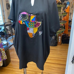 May include: Black tunic with a colorful African continent applique. The tunic has a V-neck and short sleeves. The applique features a variety of patterns and colors, including blue, yellow, red, and orange. The tunic is displayed on a mannequin.