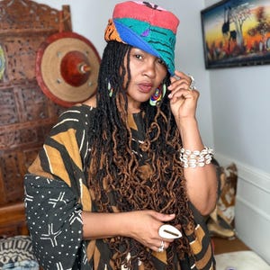 May include: A multi-colored patchwork cap featuring red, blue, green, and orange fabric panels. The cap is worn with a black and brown patterned garment, a white cowrie shell bracelet, large circular earrings, and a white ring with a gold-tone center.
