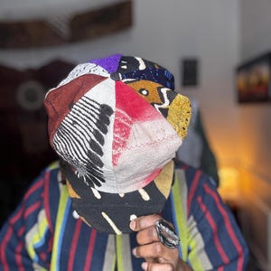 May include: A patchwork newsboy cap with a variety of fabric patterns and colors, including red, black, white, and purple. The cap is worn by a person wearing a striped shirt and a silver ring.