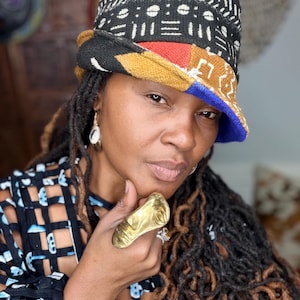 May include: A bucket hat featuring a black, white, red, and blue patterned fabric with a textured finish. The hat is paired with a large, sculptural gold-toned ring shaped like a human face and a black and blue grid-patterned top.