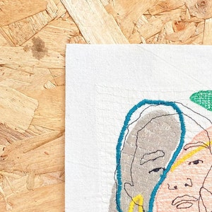 May include: Embroidered artwork featuring a stylized portrait of a person with a teal blue outline around their face. The portrait is stitched with different colored threads on a white fabric background.