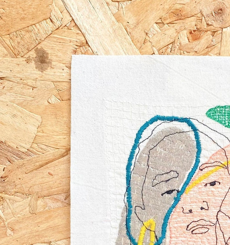 May include: Embroidered artwork featuring a stylized portrait of a person with a teal blue outline around their face. The portrait is stitched with different colored threads on a white fabric background.
