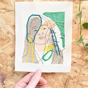 May include: Embroidered artwork featuring a stylized portrait of a person with overlapping lines and shapes in shades of green, blue, yellow, and brown.