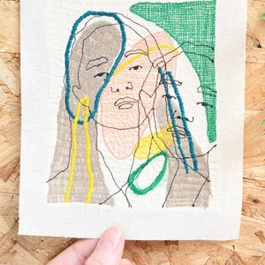 May include: Embroidered artwork featuring a stylized portrait of a person with a green, blue, yellow, and brown color scheme.