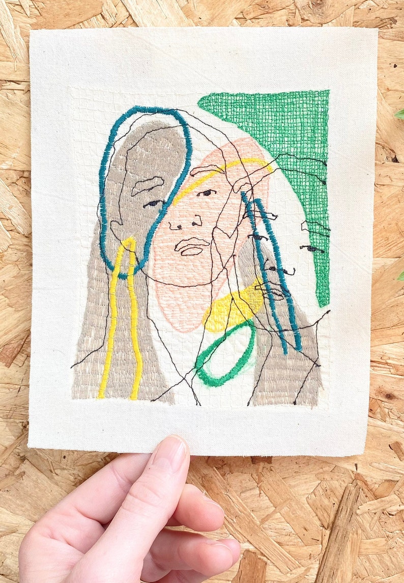 May include: Embroidered artwork featuring a stylized portrait of a person with a green, blue, yellow, and brown color scheme.