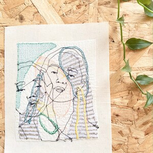 May include: A hand-stitched embroidery artwork on a beige canvas featuring a stylized portrait of a person with green, yellow, and orange thread. The artwork is framed with a white border.