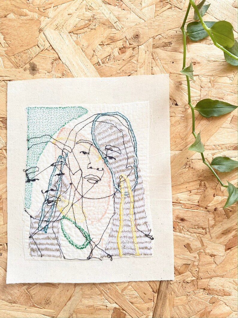 May include: A hand-stitched embroidery artwork on a beige canvas featuring a stylized portrait of a person with green, yellow, and orange thread. The artwork is framed with a white border.