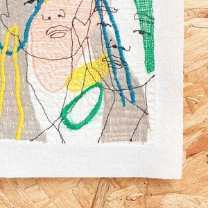 May include: A close-up of a colorful embroidered artwork featuring a face with a yellow, green, and blue design. The artwork is stitched on a white fabric.