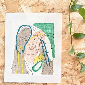 May include: Embroidered artwork on a white canvas featuring a stylized portrait of a person with overlapping lines and shapes in shades of gray, yellow, green, and blue.