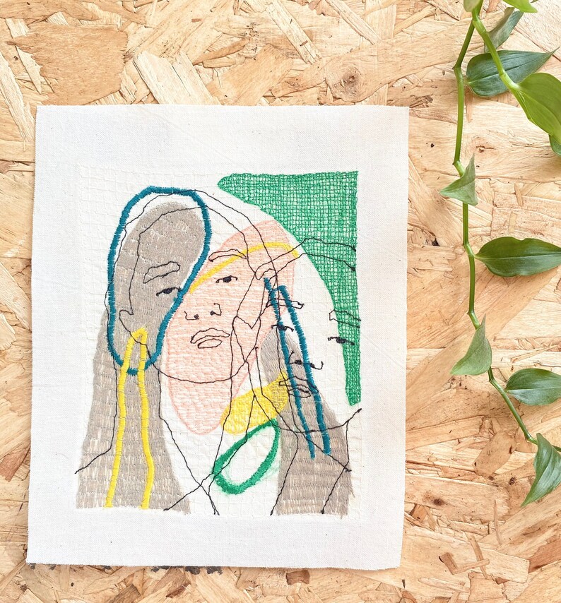 May include: Embroidered artwork on a white canvas featuring a stylized portrait of a person with overlapping lines and shapes in shades of gray, yellow, green, and blue.