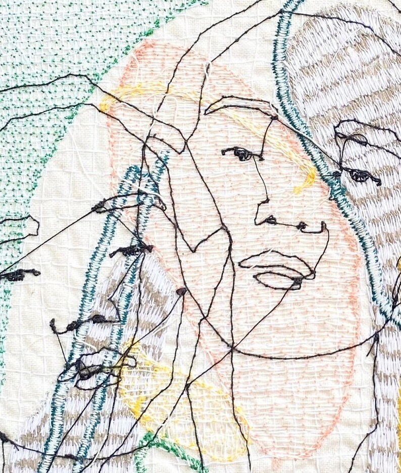 May include: A close-up of a textile art piece featuring a stylized embroidered face with multiple overlapping lines and a green, yellow, and pink color scheme.