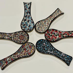 Puede incluir: Colección de seis reposacucharas de cerámica con intrincados diseños florales y geométricos en varios colores. Cada reposacucharas tiene un diseño único, incluyendo motivos florales rojos, azules y amarillos, y un borde negro. También se incluye una pieza alta en forma de jarrón.