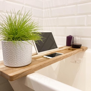 Peut inclure: Un plateau de bain en bois avec un pot blanc texturé contenant une plante verte, une tablette, un smartphone, une bougie violette et un verre à vin avec du vin rouge. Le plateau est en bois clair.