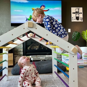 May include: A white wooden climbing structure with colorful rungs and wooden dowels. Two children are interacting with the structure. One child is climbing on the structure, while the other is sitting on the floor. The background includes a TV and artwork.