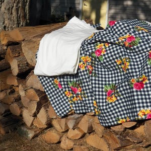 May include: A stack of firewood with a white sheet and a black and white checkered fabric with floral patterns. The fabric has red and yellow roses. The scene is outdoors, with a tree and a building in the background.