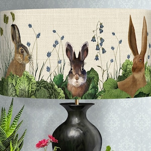 May include: A black lamp with a cream lampshade featuring a whimsical design of three brown rabbits peeking out from behind green leafy plants and blue flowers.