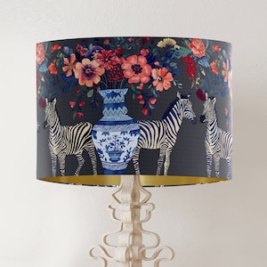 May include: A lampshade with a dark grey background featuring a floral pattern with pink, red, and white flowers. Three zebras are depicted in the pattern, along with a blue and white porcelain vase.