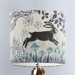 May include: A cream-colored lampshade with a blue and black illustration of a rabbit running through a forest. The illustration features stylized trees, flowers, and mushrooms.