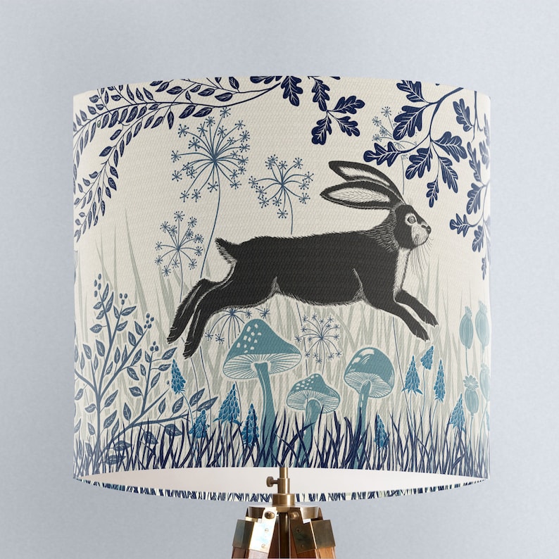 May include: A cream-colored lampshade with a blue and black illustration of a rabbit running through a forest. The illustration features stylized trees, flowers, and mushrooms.
