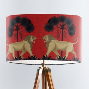 May include: A red lampshade with a black and gold design featuring two lions facing each other in a jungle setting. The lampshade is on a wooden tripod stand.