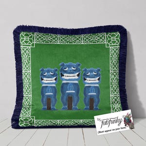 May include: Square cushion with a green centre featuring three blue cartoon figures. The cushion has a white geometric border, a navy blue fringe, and a tag that says "fabfunky".