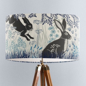 May include: A lampshade with a white background and a blue and black floral pattern featuring two black rabbits. The rabbits are in a field of flowers and grass.