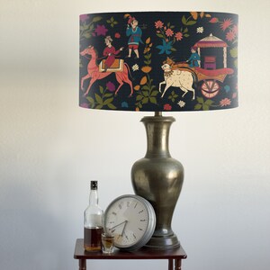 May include: A black lampshade with a colorful Indian-inspired design featuring people riding animals and a carriage. The lampshade is on a lamp with a gold-colored base.